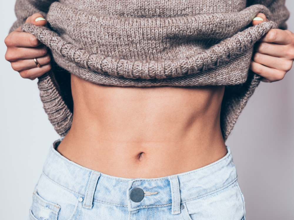 Close-up of flat female stomach. Stylish young woman taking off warm knitted sweater showing her belly and waist.